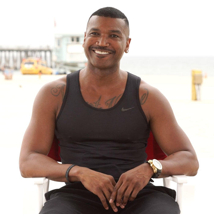 Image of Kendall Hogan, a smiling black man is sitting in a chair on the beach. He wears a black tank top.