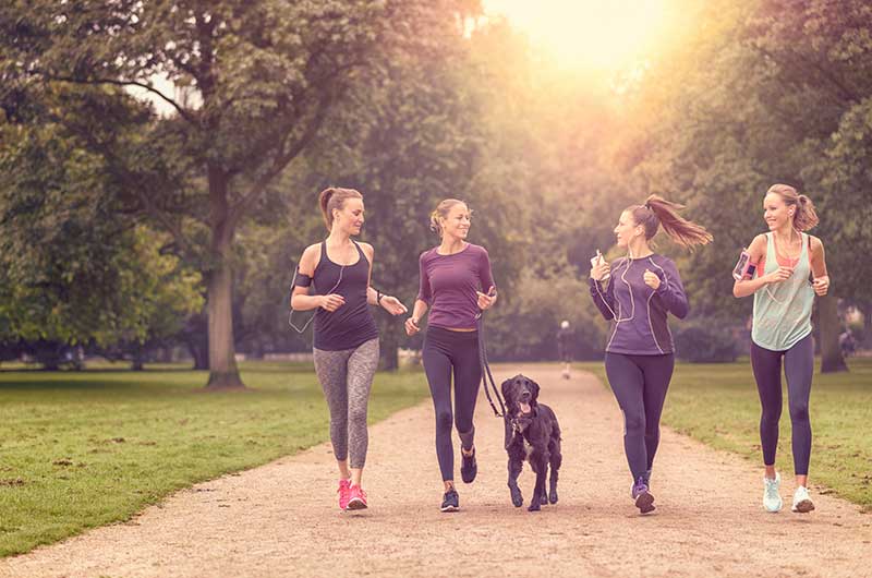 Four women and a black dog walking toward the camera down a dirt path lined by grass and tall trees in the daytime. All the women are smiling and wearing dark exercise pants and brightly colored tops.